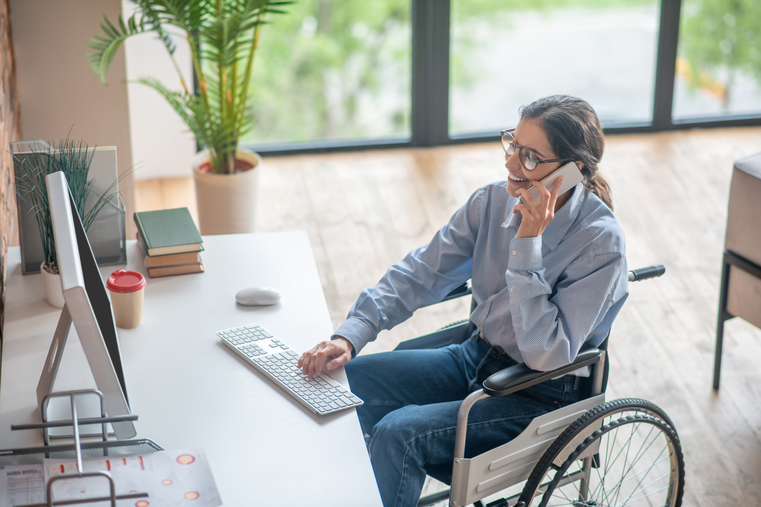 A young woman in a wheelchair is talking on the phone at her office desk